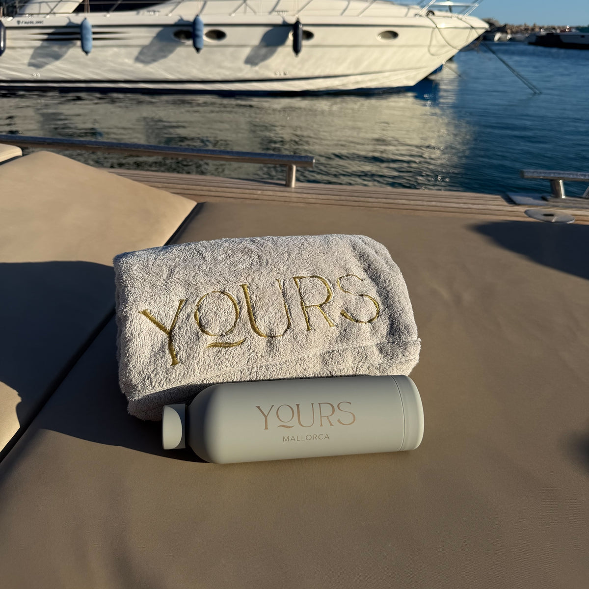 The YOURS bottle beside the embroidered YOURS towel on a sun lounger aboard